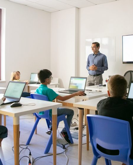 Confident teacher explaining lesson to pupils. Multiethnic children sitting at table in classroom, listening middle-aged man and using laptop computers. Childhood and digital education concept